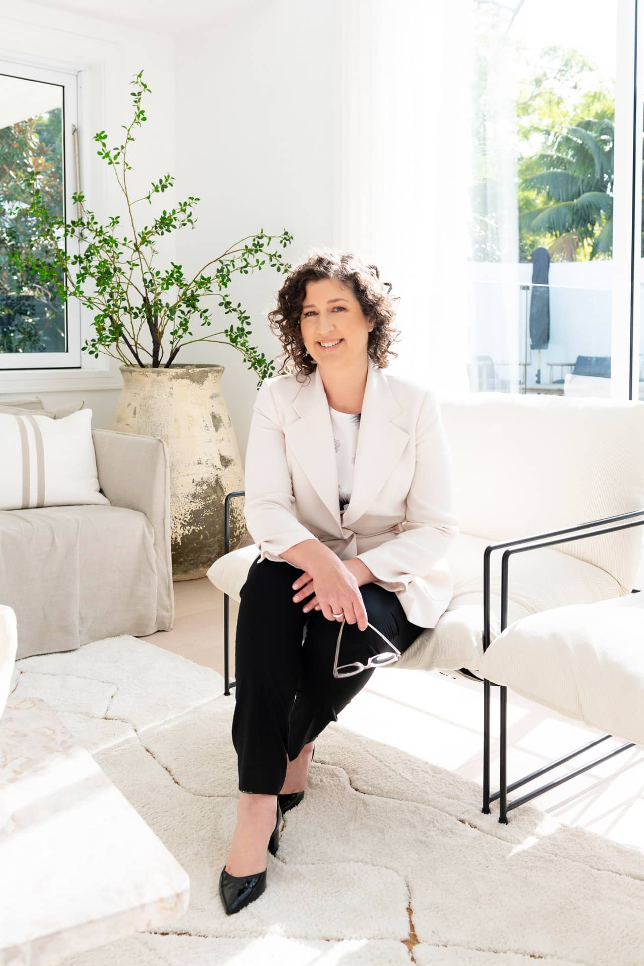 Marnie Cooper sits on an off-white sofa in a sunlit room in Sydney, holding her glasses. She wears a light blazer and dark pants, with a large plant in the background—a tranquil setting perfect for meditation.